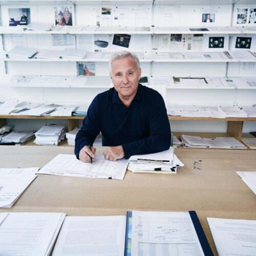Ian Schrager in a navy sweater seated at a wooden desk covered in papers, shelves of documents behind him.