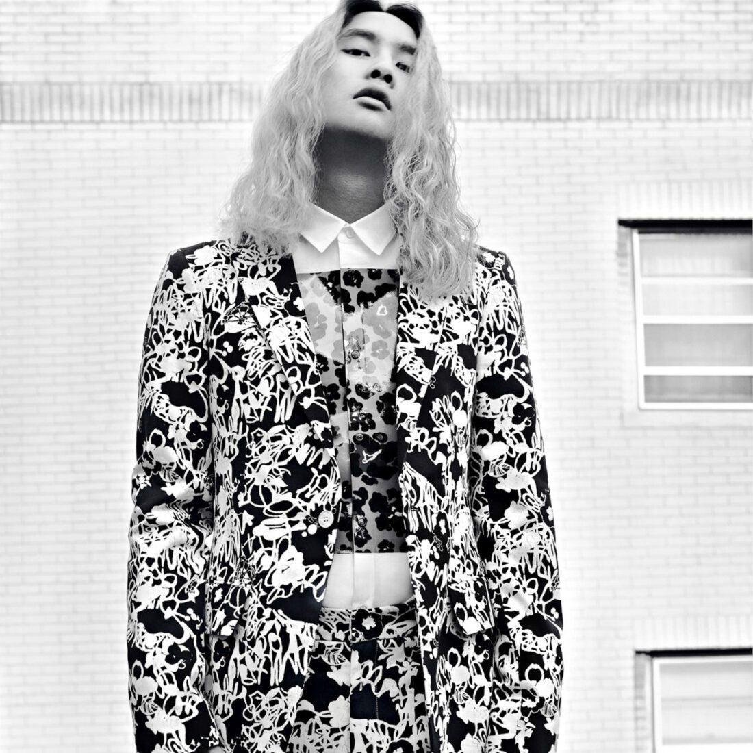Black and white portrait of Korakrit Arunanondchai in a floral print suit before a white brick wall.