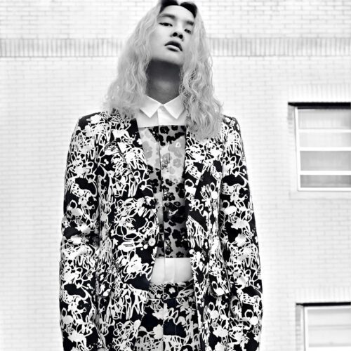 Black and white portrait of Korakrit Arunanondchai in a floral print suit before a white brick wall.