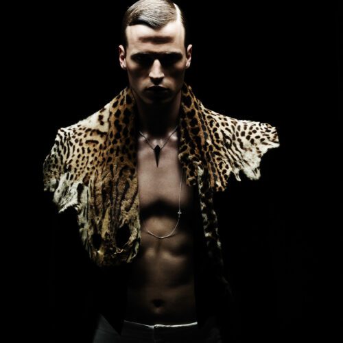 Lars Burmeister shirtless with a leopard-print pelt over his shoulders and pendant necklaces, lit against black.