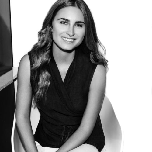Black-and-white portrait of Lauren Bush smiling, seated in a white chair, wearing a sleeveless dark wrap top with long wavy hair.