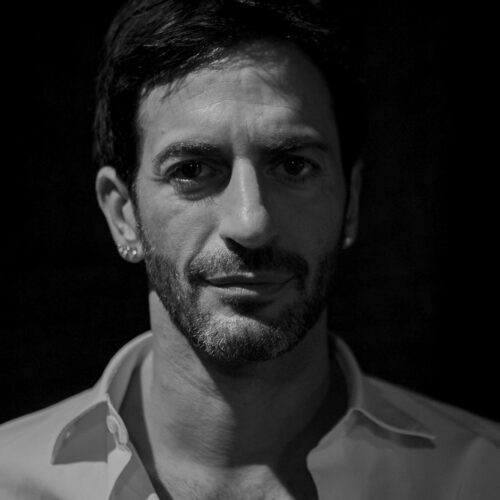 Black and white close-up portrait of Marc Jacobs with stubble and open-collared shirt against a dark background.