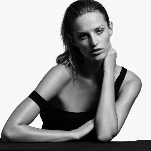 Black-and-white portrait of Michaela D with slicked-back hair, hand at her neck, in a black off-shoulder top.