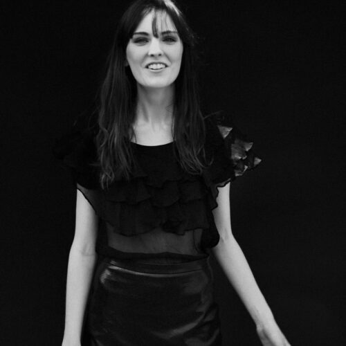 Black-and-white portrait of Myf Shephard smiling in a sheer ruffled black blouse and leather skirt against a dark backdrop.