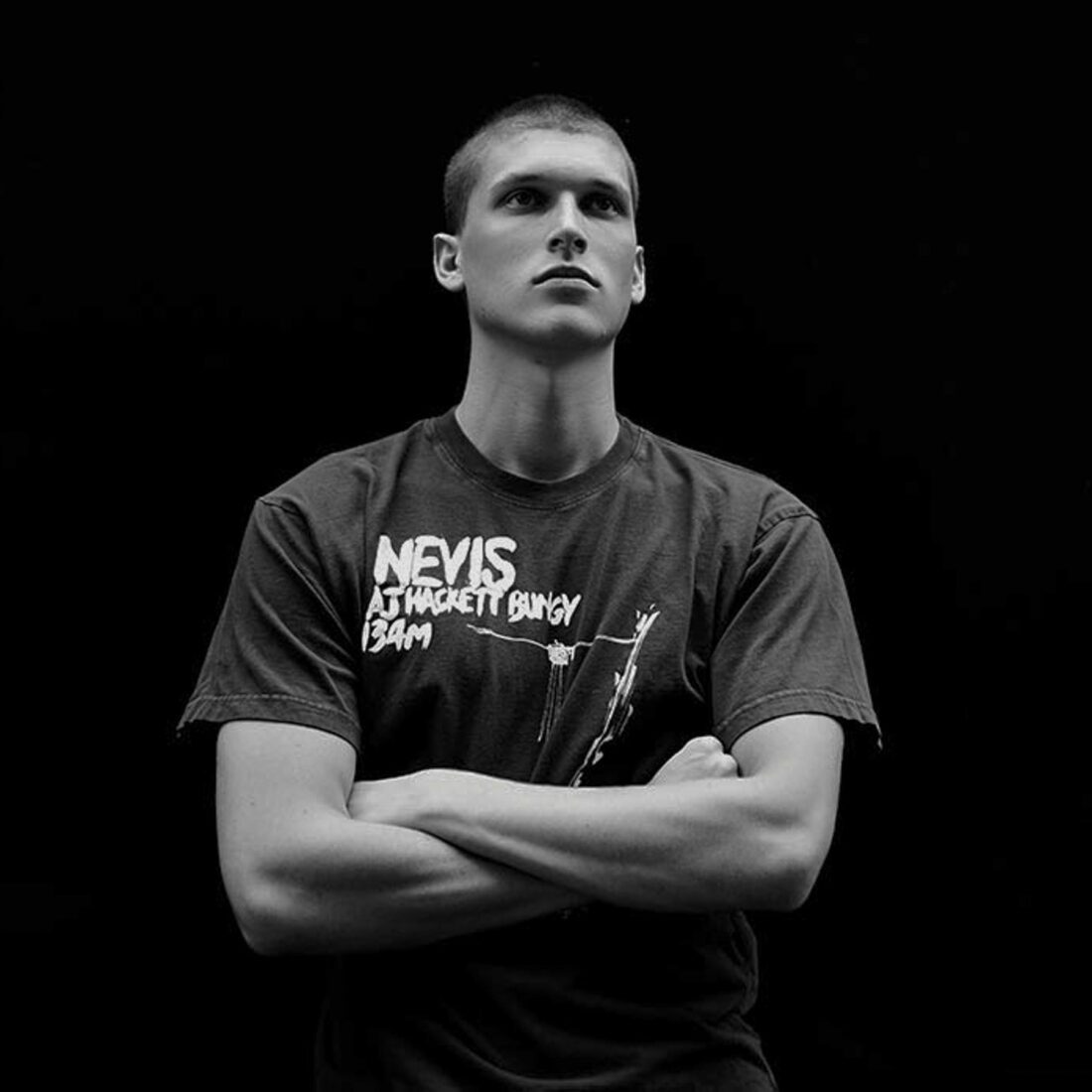 Black and white portrait of a young man with a buzz cut, arms crossed, wearing an AJ Hackett Nevis Bungy T-shirt.
