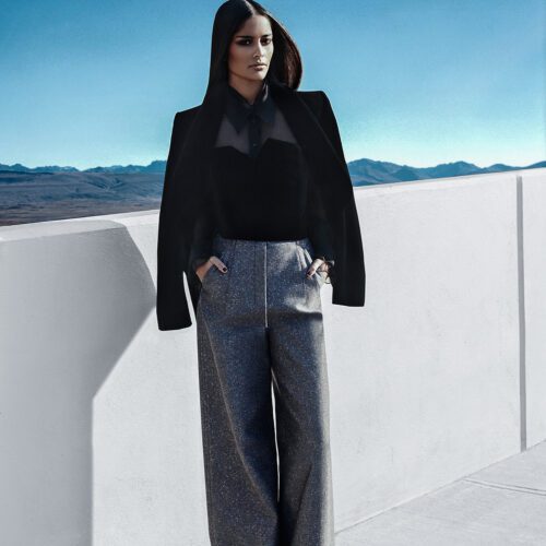Model in black blouse, draped jacket and grey wide-leg trousers on a white rooftop above mountains, for The New York Times.
