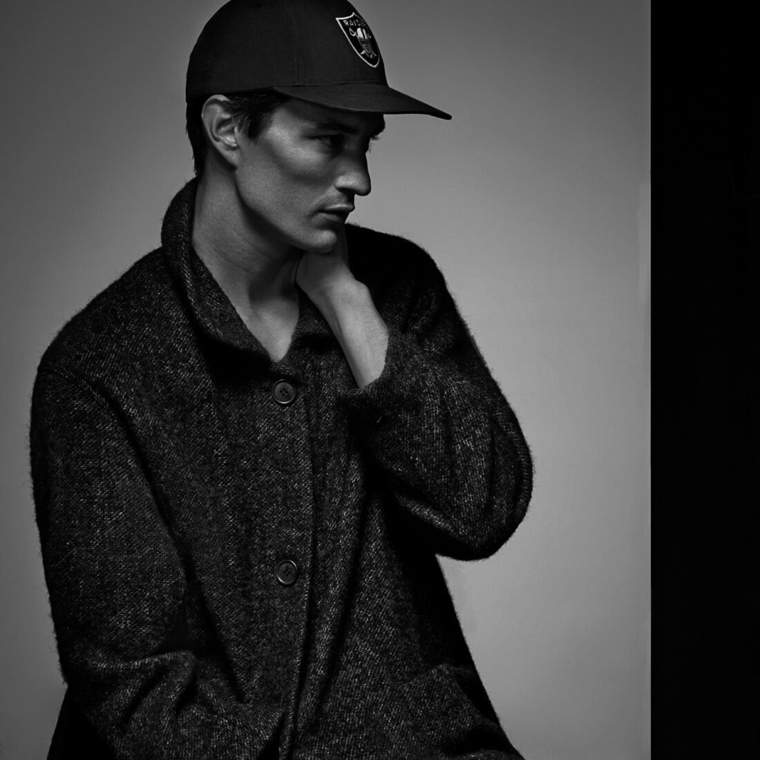 Black-and-white portrait of Owen Stuart in a Raiders cap and textured wool coat, hand at his neck, looking aside.