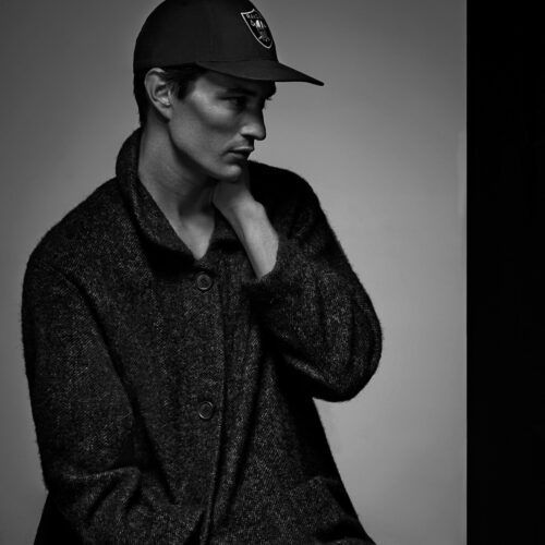 Black-and-white portrait of Owen Stuart in a Raiders cap and textured wool coat, hand at his neck, looking aside.