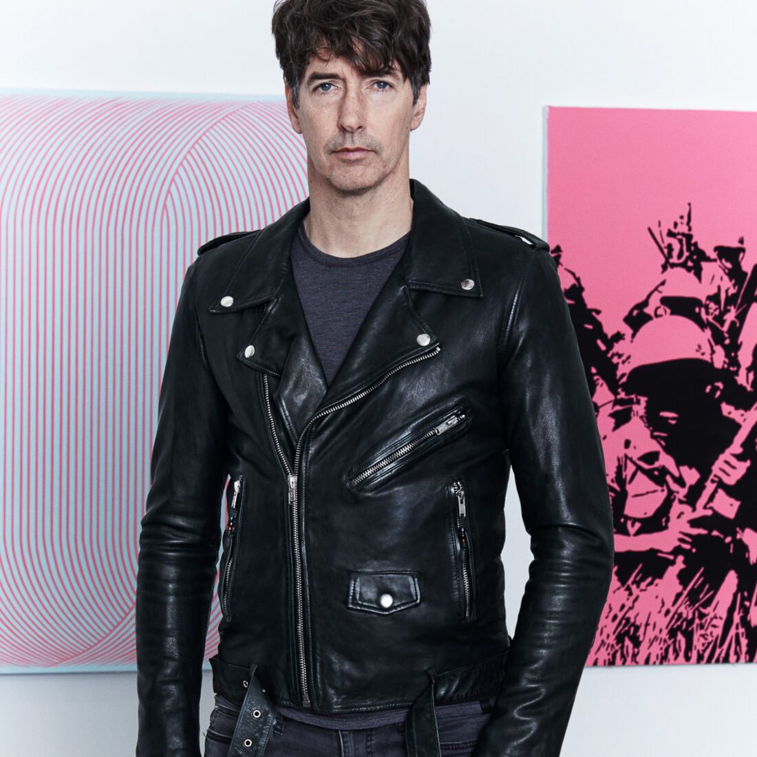 Richard Phillips in a black leather biker jacket standing before pink op-art and pop-art canvases.