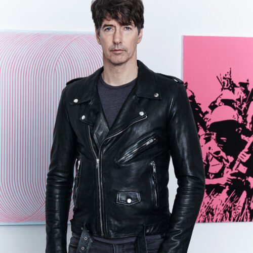 Richard Phillips in a black leather biker jacket standing before pink op-art and pop-art canvases.