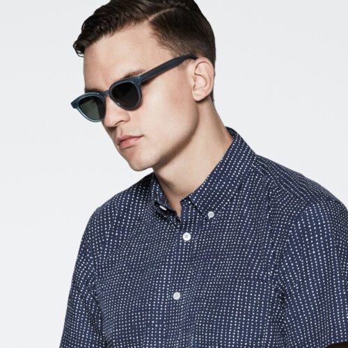 Man in blue-framed sunglasses and a navy short-sleeve shirt with white dots against a pale grey backdrop, for Saturdays NYC.