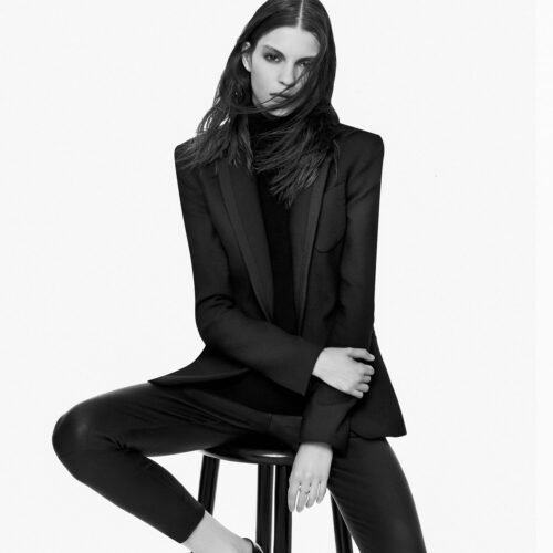 Black and white shot of a model in a black blazer, turtleneck and leather leggings seated on a stool, for Theory.