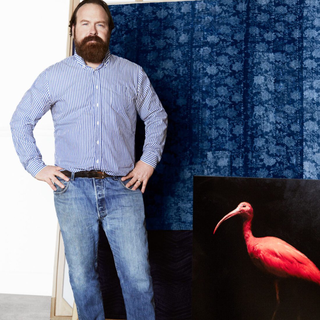 Todd Murphy in a striped shirt and jeans, hands on hips, beside a blue damask backdrop and a painting of a scarlet ibis.
