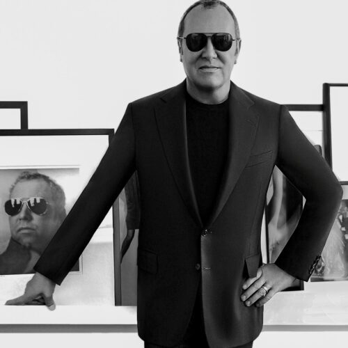 Black and white portrait of Michael Kors in sunglasses and a dark suit before framed photographs, for Vogue China.