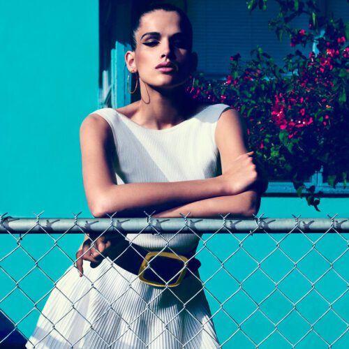 Model in white ribbed knit dress with wide gold-buckle belt leaning on a chain-link fence before a turquoise wall, for Amica.