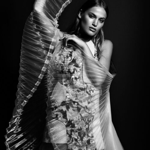 Black-and-white portrait of Sonya Garalova holding sheer pleated fabric over an embellished gown against a dark backdrop.