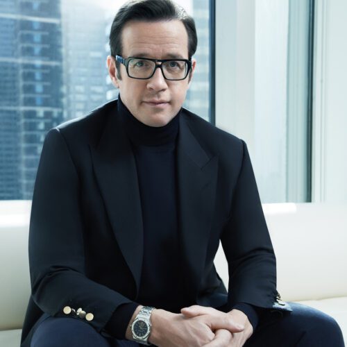 François-Henry Bennahmias in glasses, black suit and turtleneck, seated on a white sofa by a city window, for Audemars Piguet.