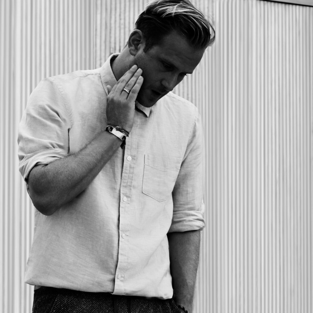 Black and white shot of a man in a rolled-sleeve linen shirt touching his face before a ribbed metal wall, for Hercules.