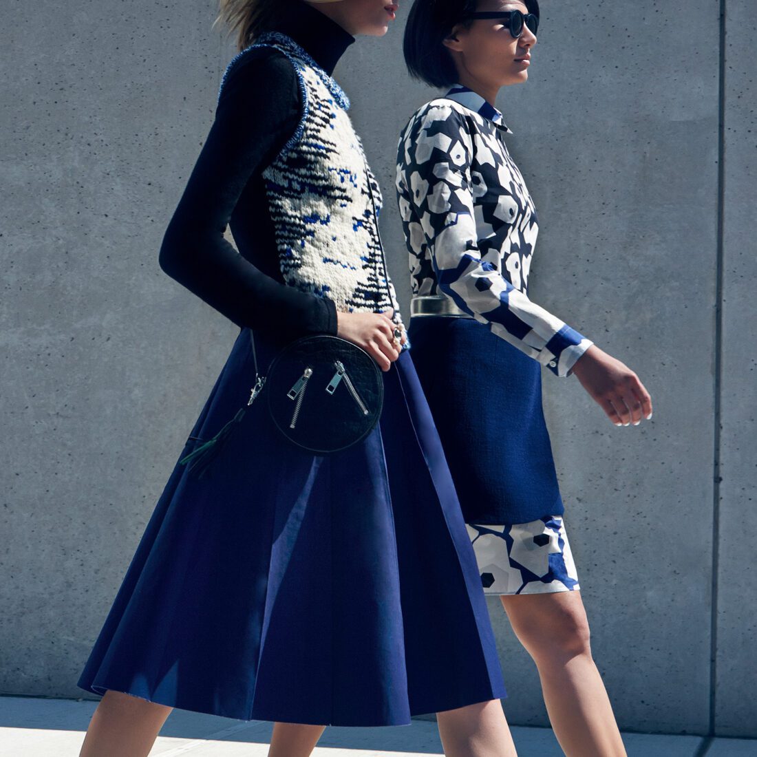 Two models walk past a concrete wall in navy skirts, a knit vest and a blue-and-white print dress, for InStyle.