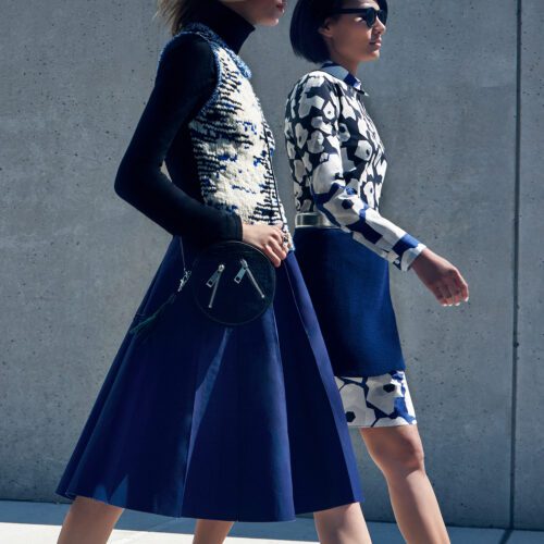 Two models walk past a concrete wall in navy skirts, a knit vest and a blue-and-white print dress, for InStyle.