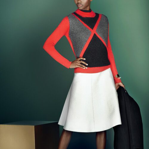 Model in a red and grey argyle-panel sweater and white A-line skirt holding a black coat against a green backdrop, for InStyle.