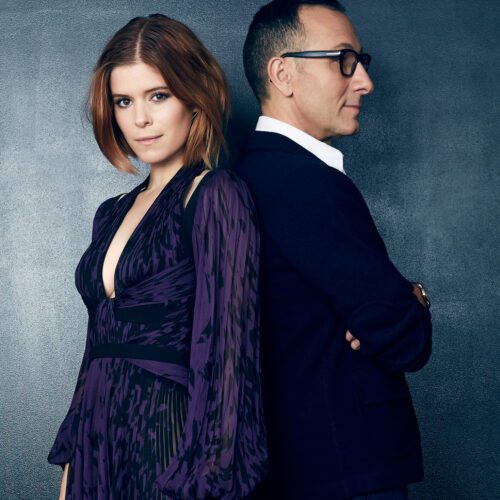 Kate Mara in a purple print dress standing back to back with Gilles Mendel in a navy blazer against a grey wall.