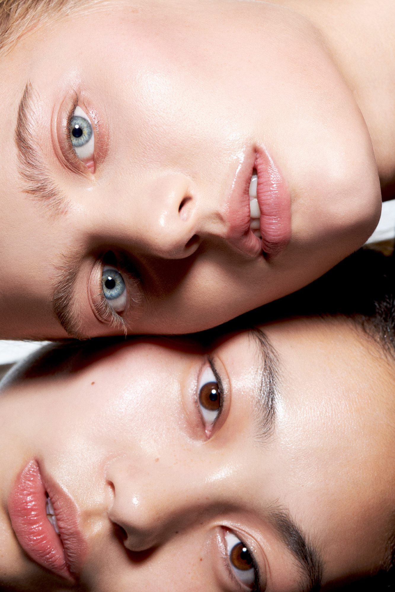 Close-up beauty shot of two models lying head to head, one blue-eyed and one brown-eyed, with glossy pink lips, for Buro.