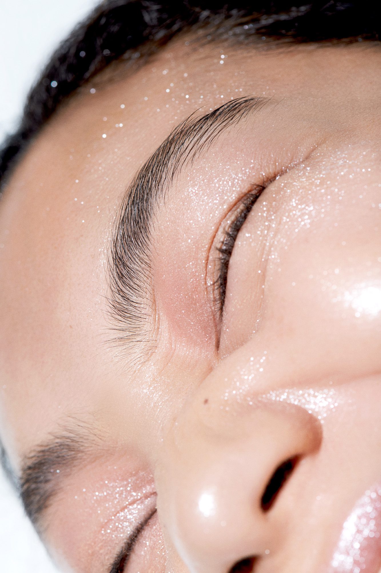 Close-up of a face lying sideways with eyes closed, pink eyeshadow and fine glitter on dewy skin, for Buro.