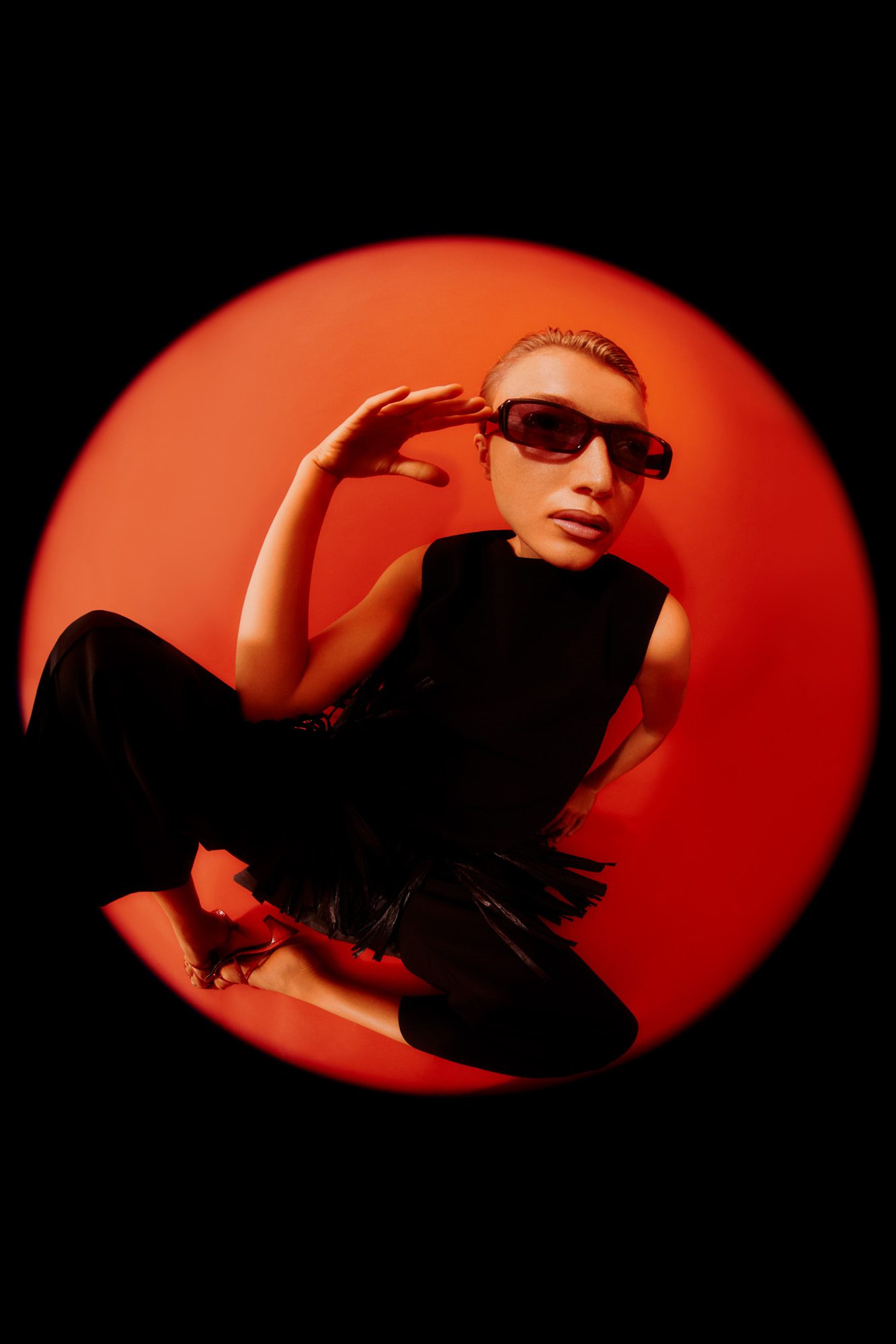 Model in black sunglasses and a black fringed dress crouching inside a red circle of light on black, for Numéro.