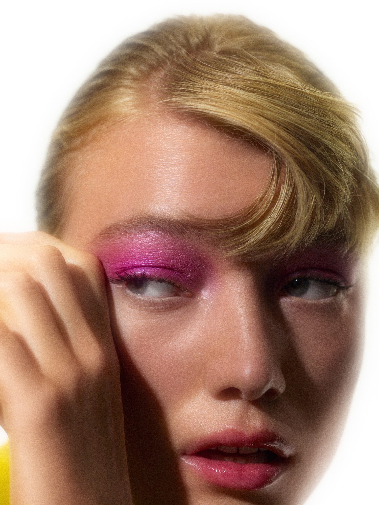 Close-up of a blonde model with magenta eyeshadow and glossy pink lips, hand pressed to her temple, for Numéro.