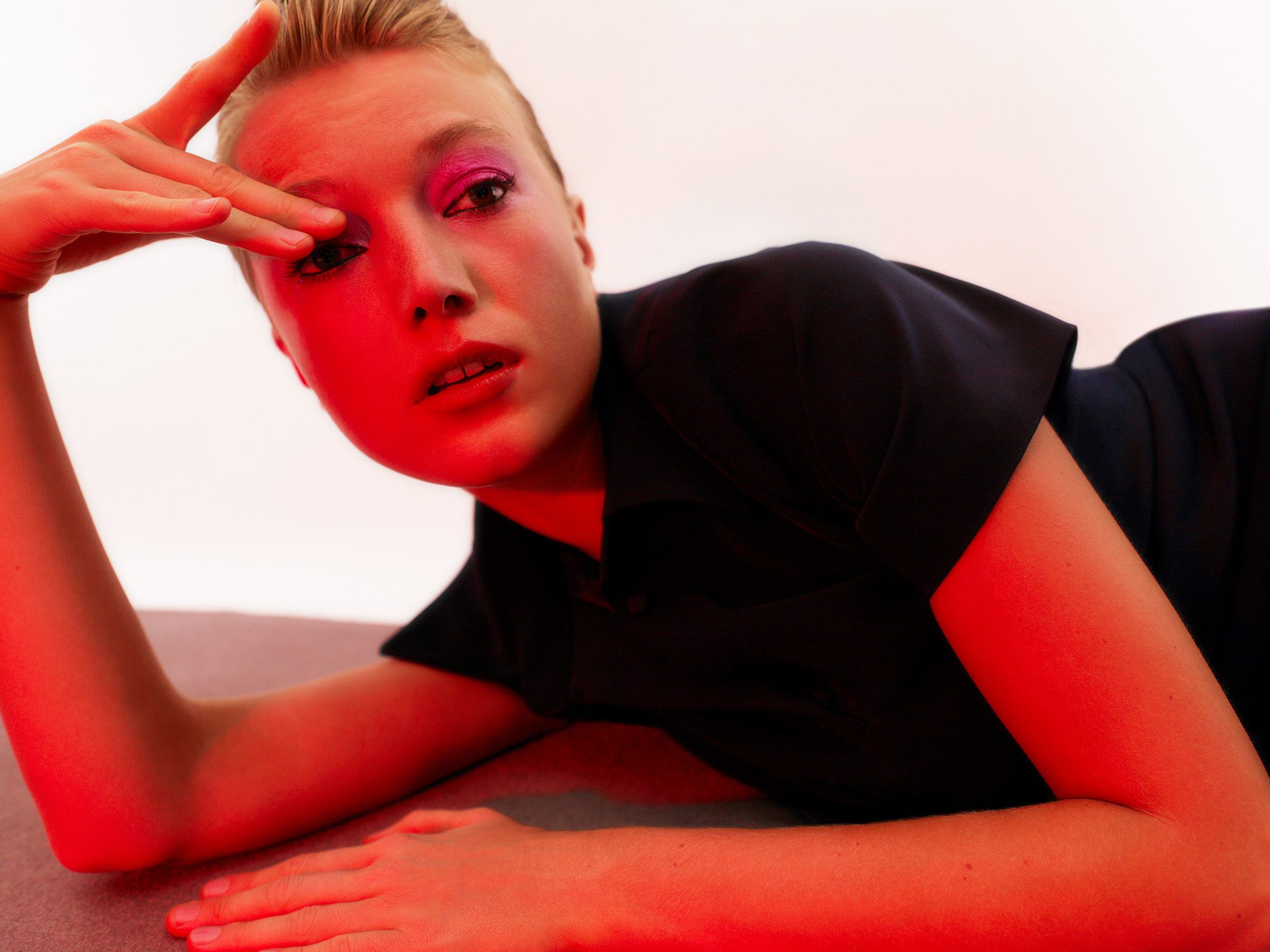 Model with short blonde hair and pink eyeshadow lying on the floor in a black top, bathed in red light, for Numéro.