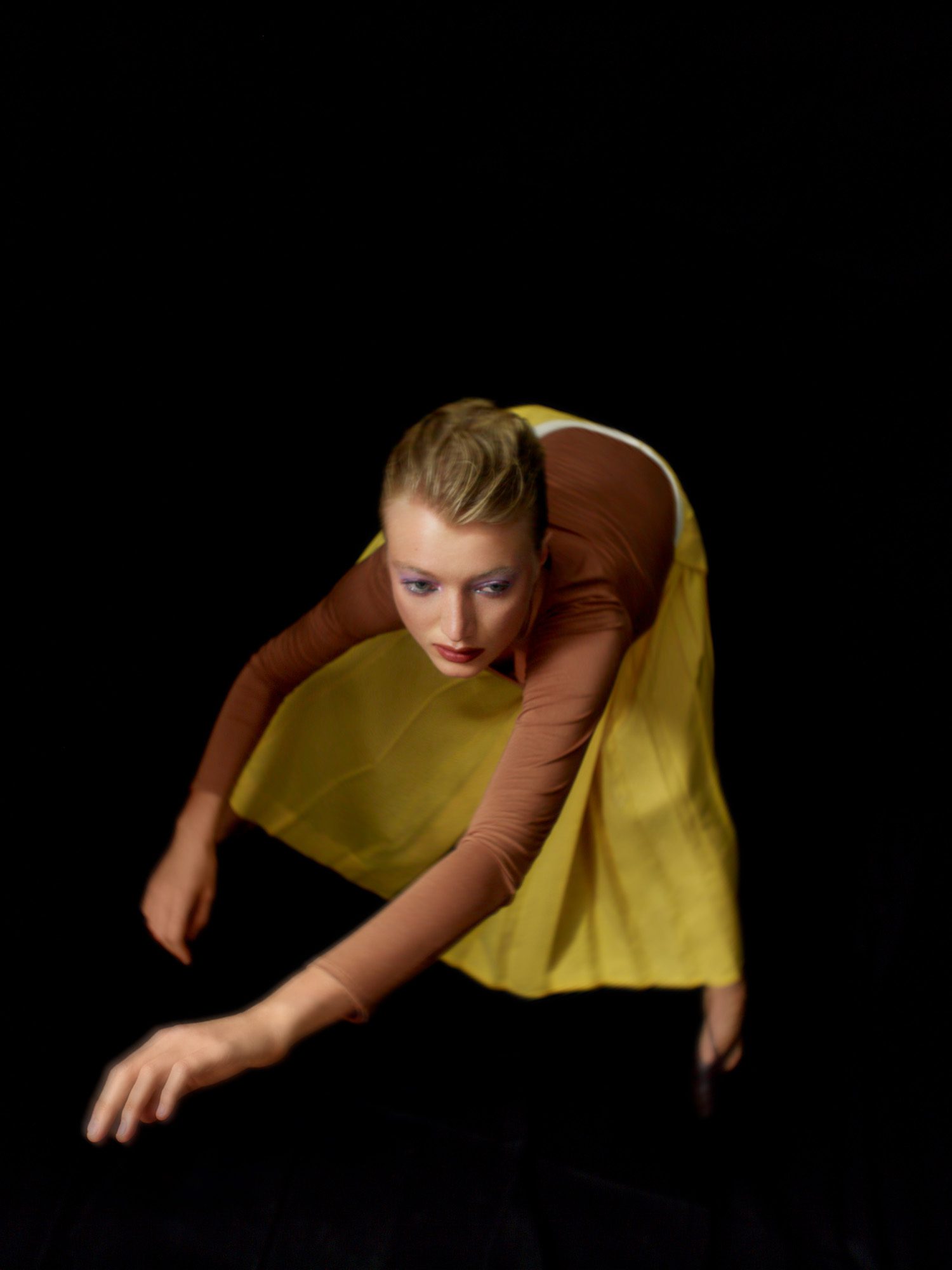 Blonde model leaning forward in a rust long-sleeve top and yellow silk skirt against a black background, for Numéro.