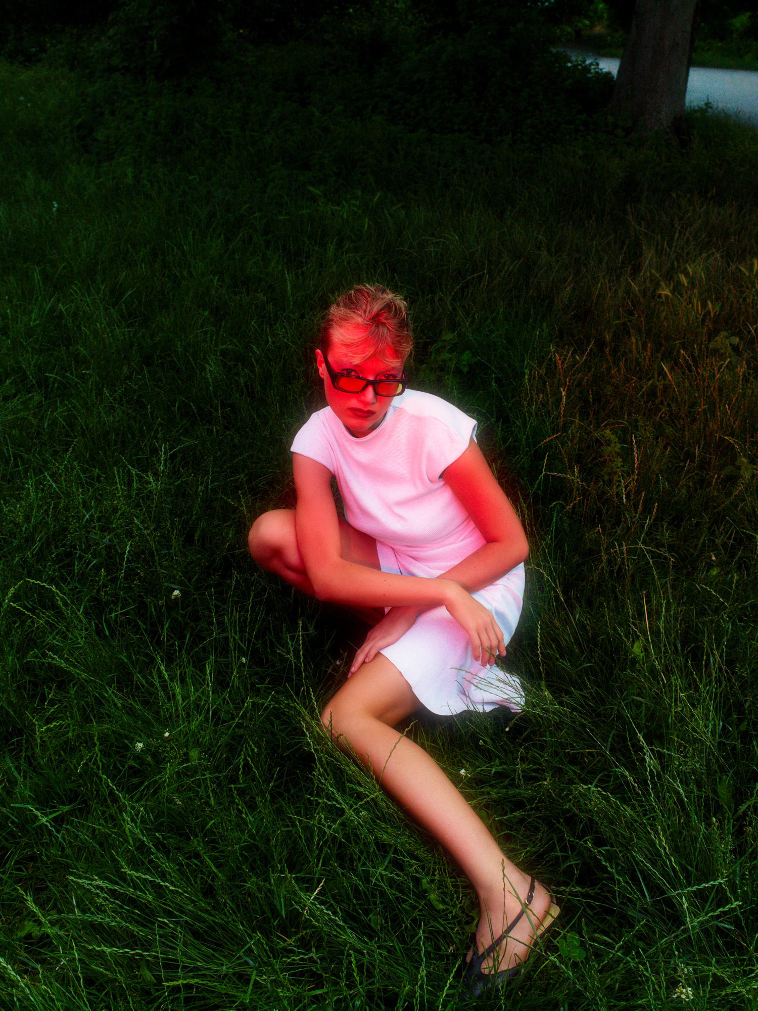 Model in a white dress, tinted sunglasses and sandals crouching in tall dark grass at dusk, lit by flash, for Numéro.