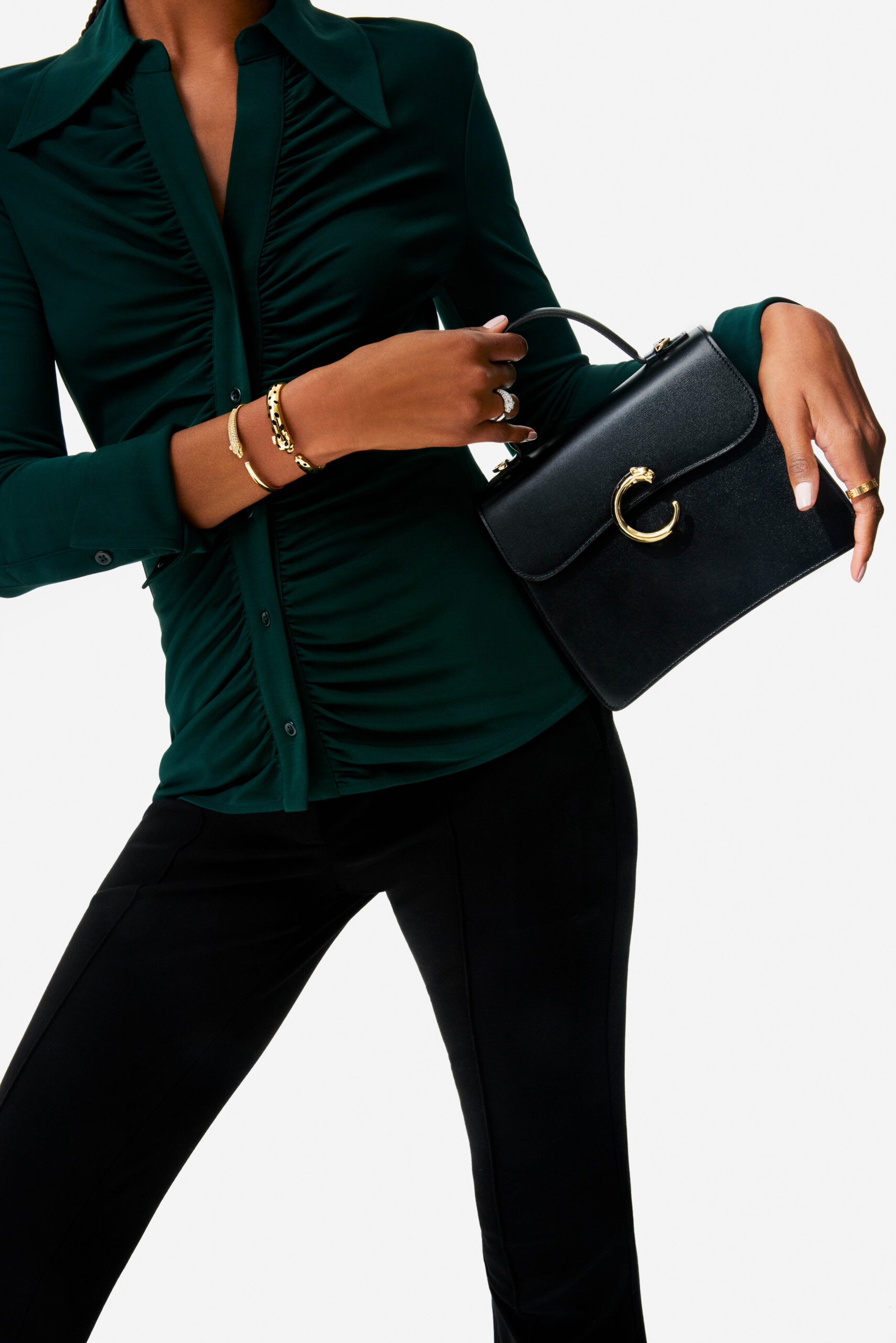 Model in a ruched green shirt and black trousers holding a black bag with gold panther clasp and gold bracelets, for Cartier.