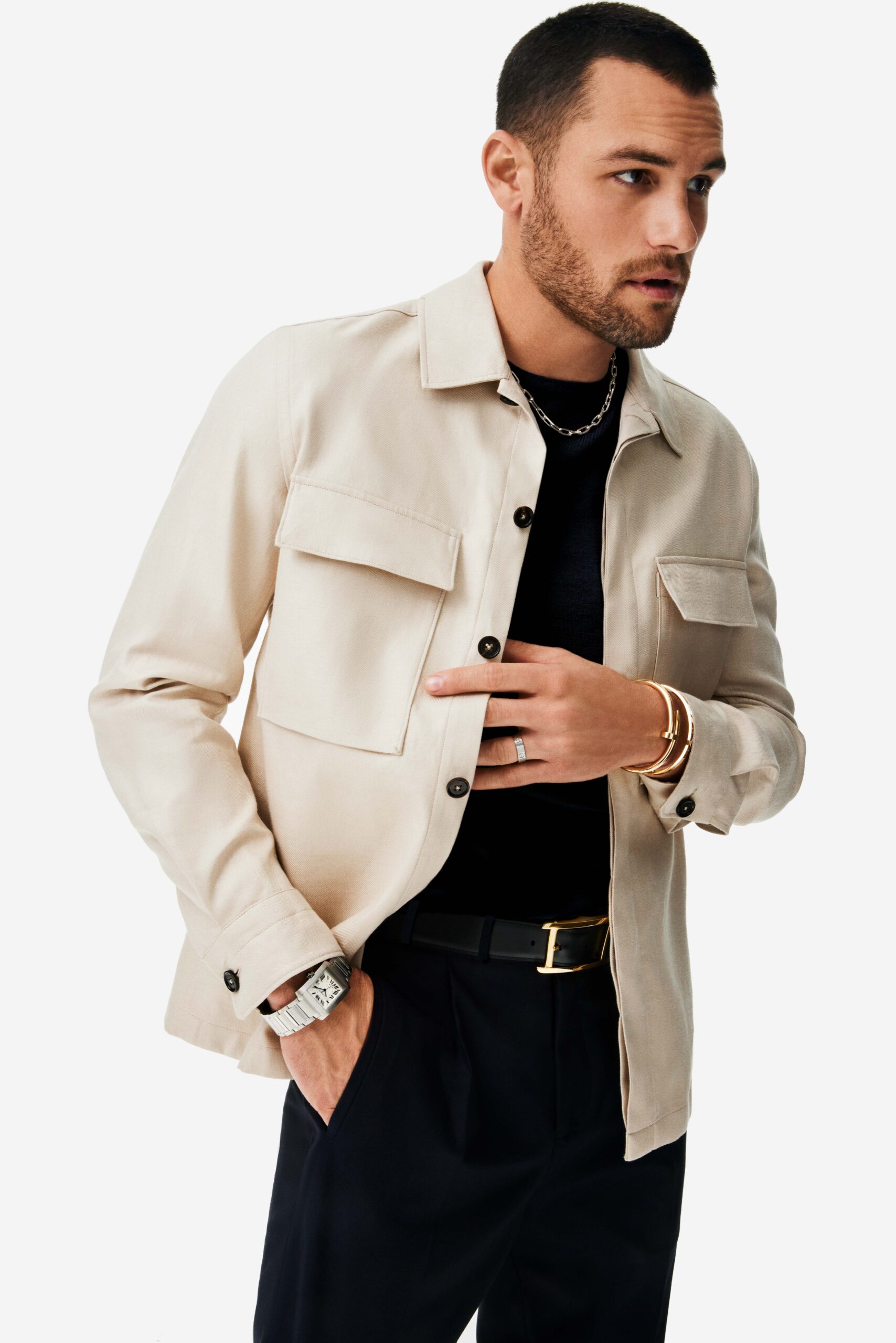 Man in a cream overshirt and black tee wearing a steel watch, gold bracelets and a silver chain, for Cartier.