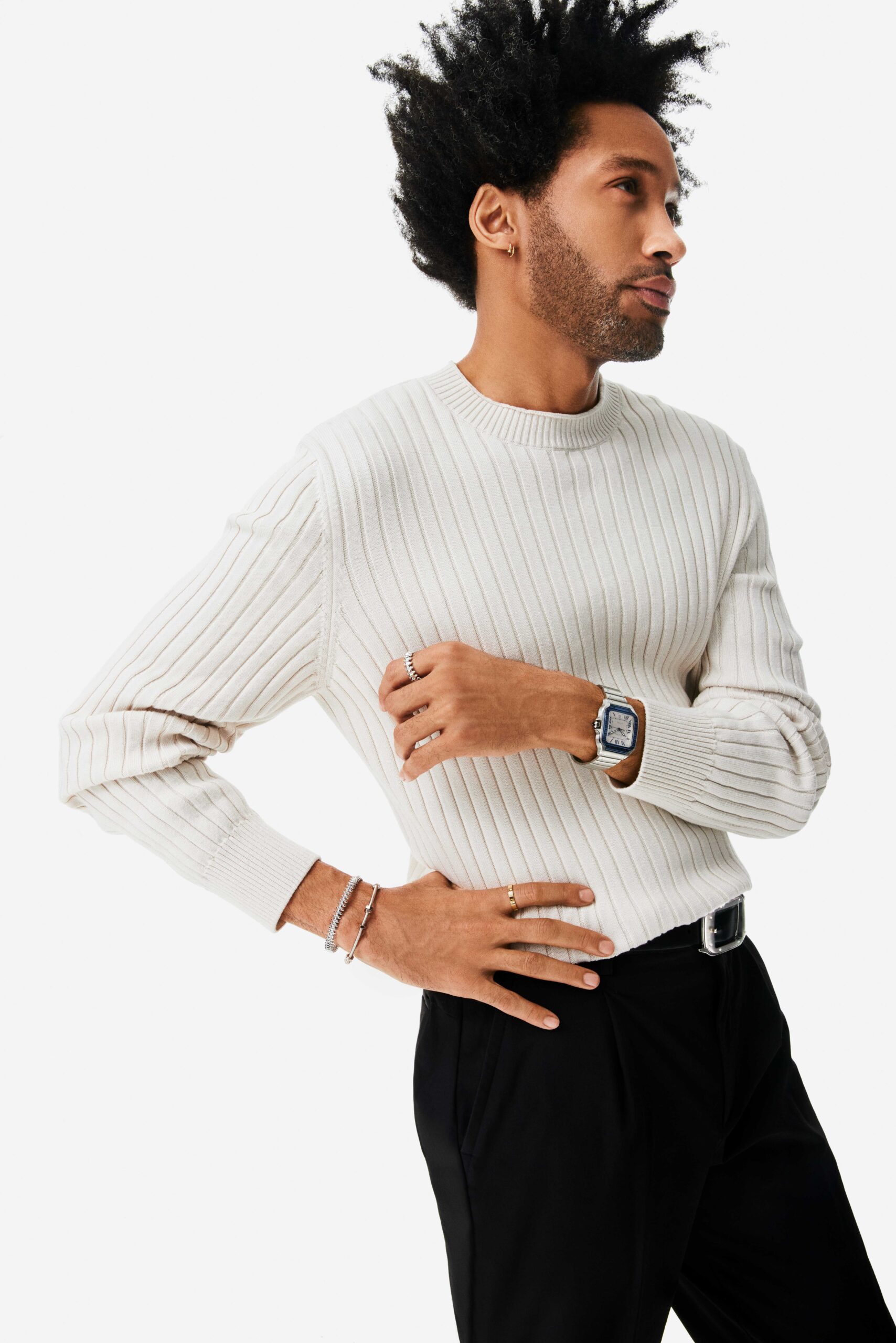 Man in a cream ribbed sweater and black trousers wearing a steel square watch, rings and bracelets, for Cartier.