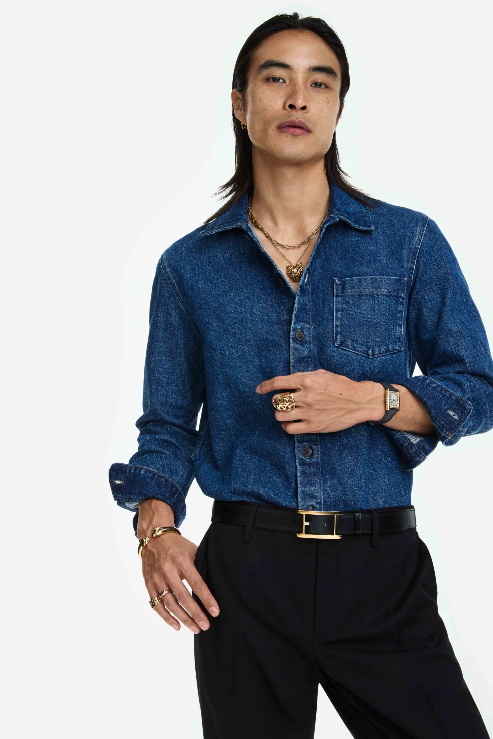 Model in a blue denim shirt and black trousers wearing gold necklaces, rings, bracelet and a Tank watch, for Cartier.