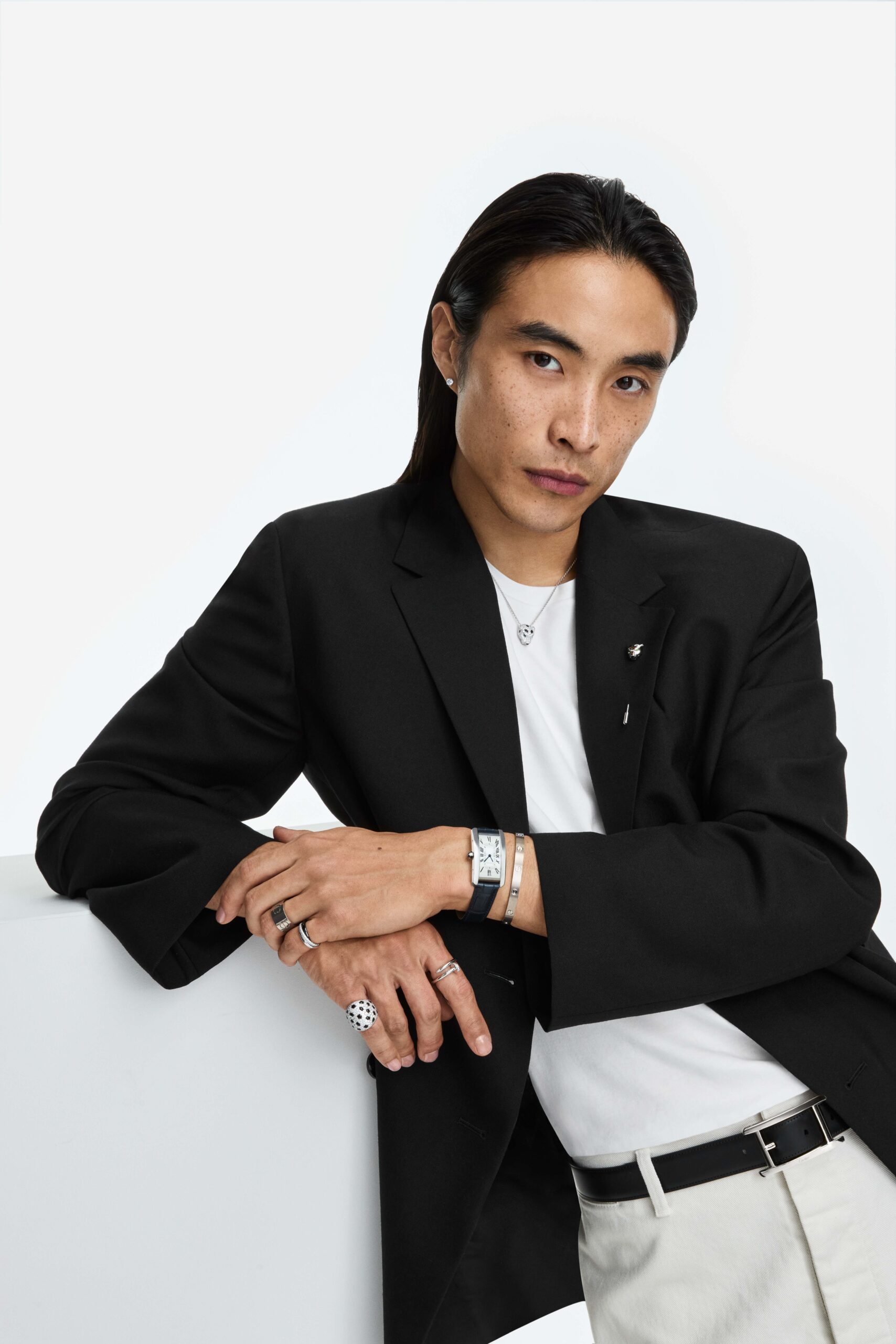 Man with long dark hair in a black blazer and white tee, leaning on a white block, wearing a watch, bracelet and rings, for Cartier.