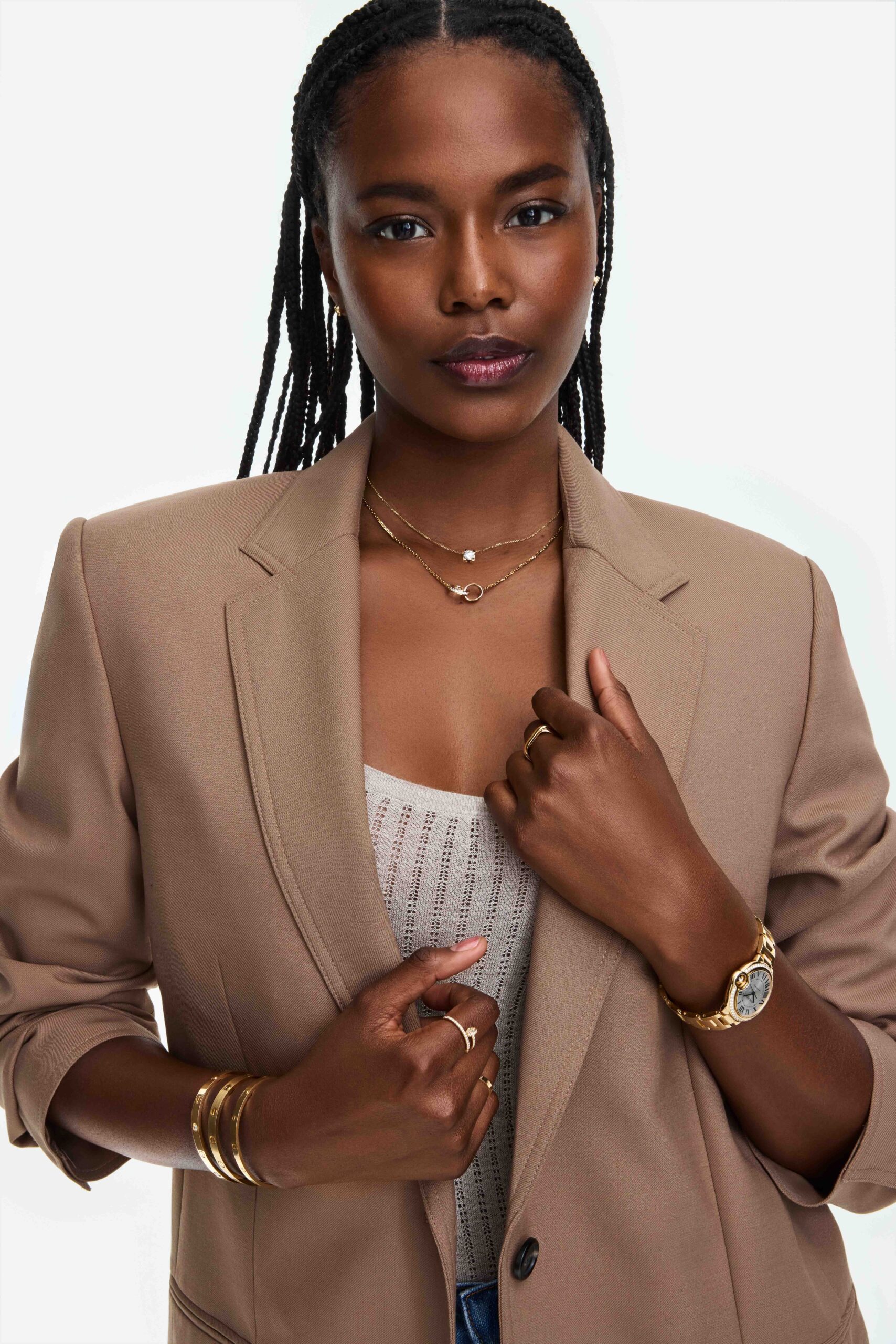 Model in a camel blazer and knit top wearing gold necklaces, rings, bangles and a gold watch on white, for Cartier.