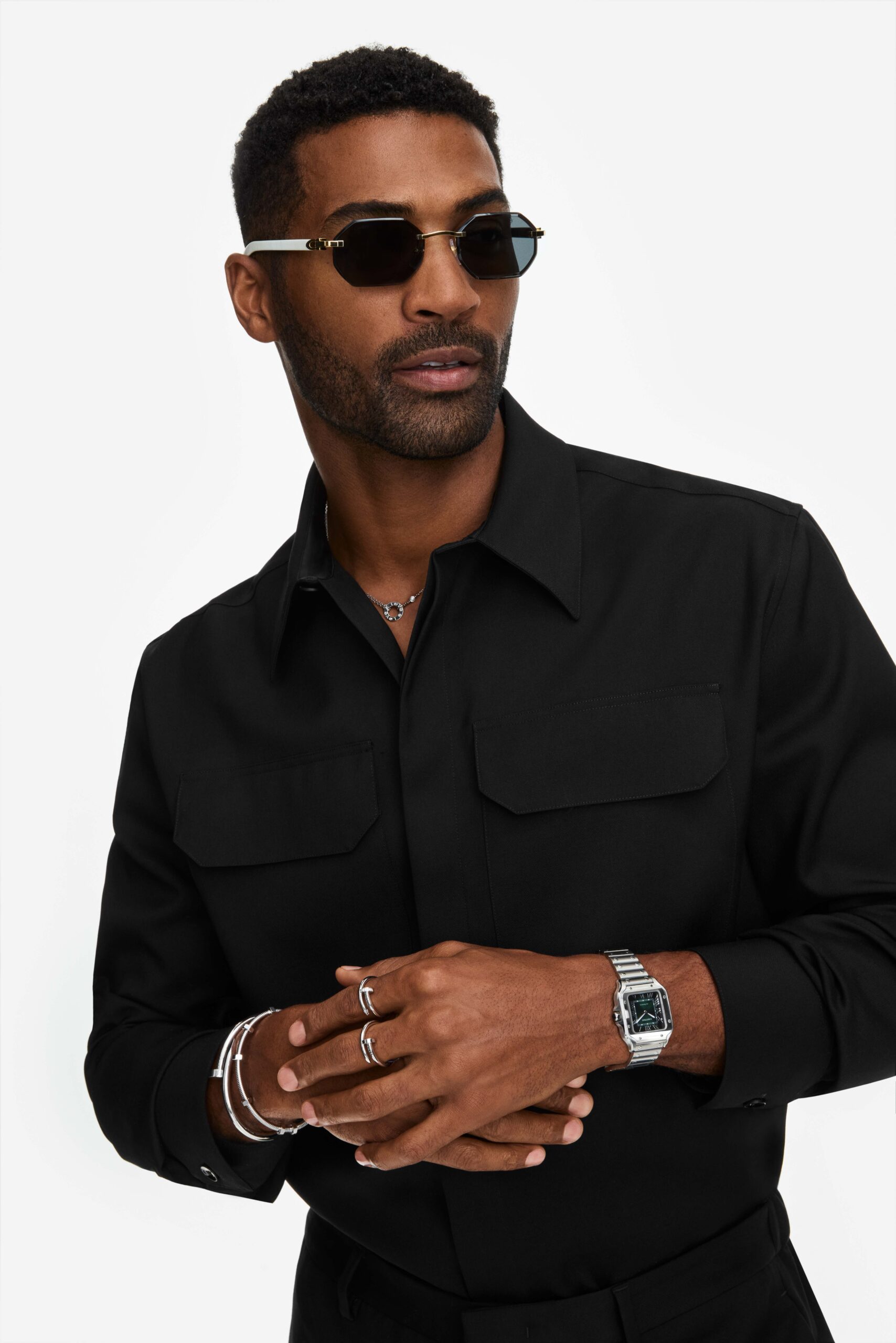 Man in black shirt and octagonal sunglasses wearing a steel watch, silver bracelets and rings, for Cartier.