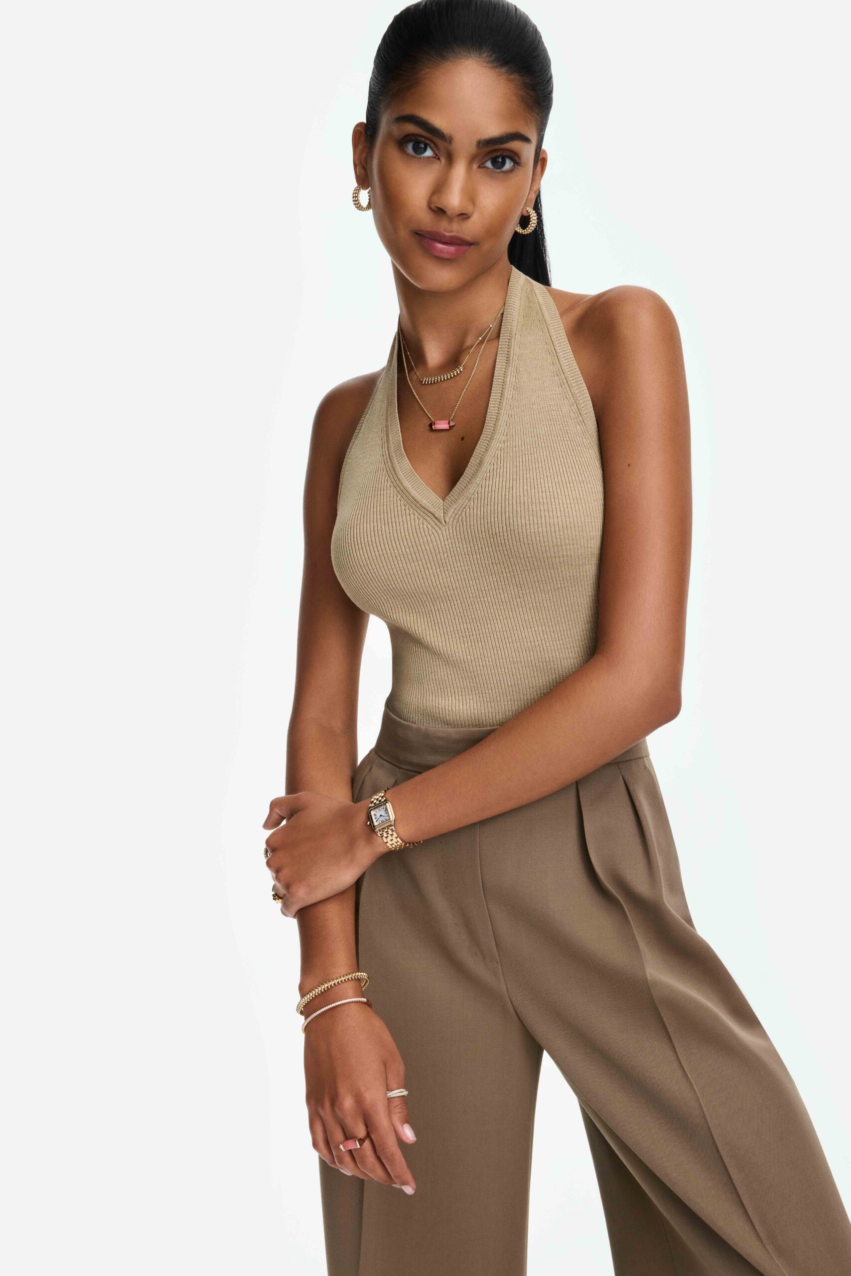 Model in a beige knit halter top and taupe trousers wearing a gold watch, necklaces and bracelets, for Cartier.