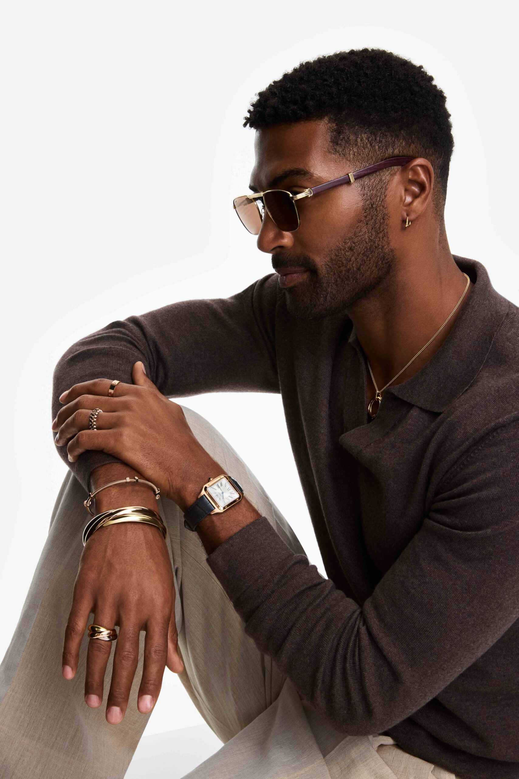Man in brown knit polo and tinted sunglasses wearing gold bracelets, rings and a square watch, seated on white, for Cartier.