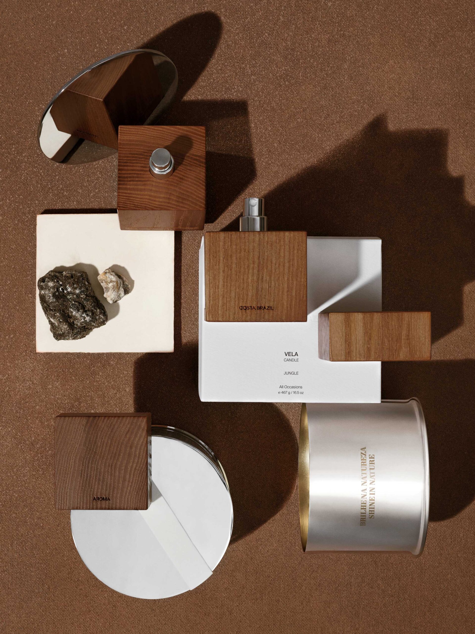 Still life of wood-capped fragrance bottles, Vela candle box, silver tin and stones on brown ground, for Costa Brazil.