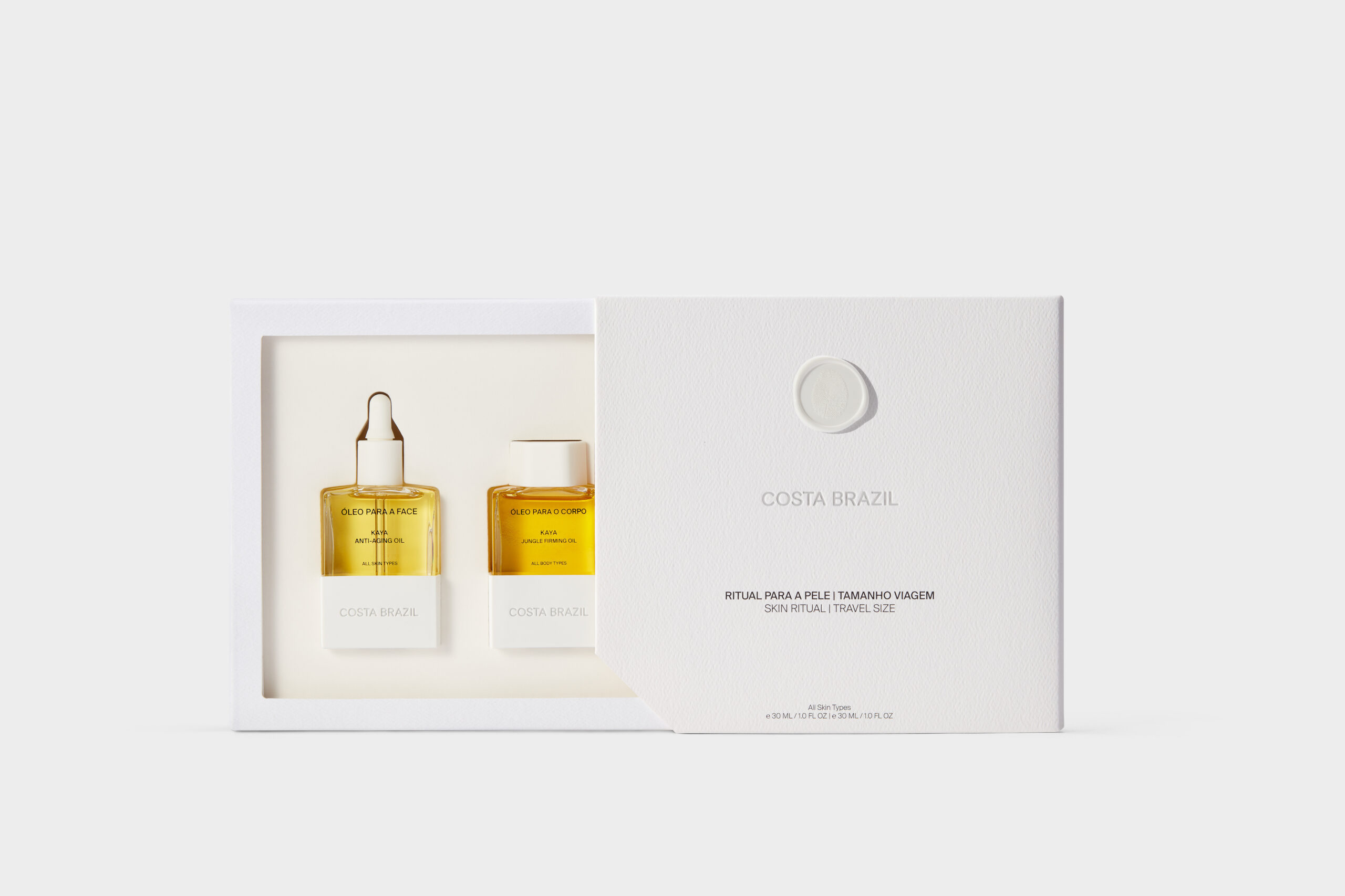 Still life of a white gift box opened to show face and body oil bottles in golden oil on a grey backdrop, for Costa Brazil.