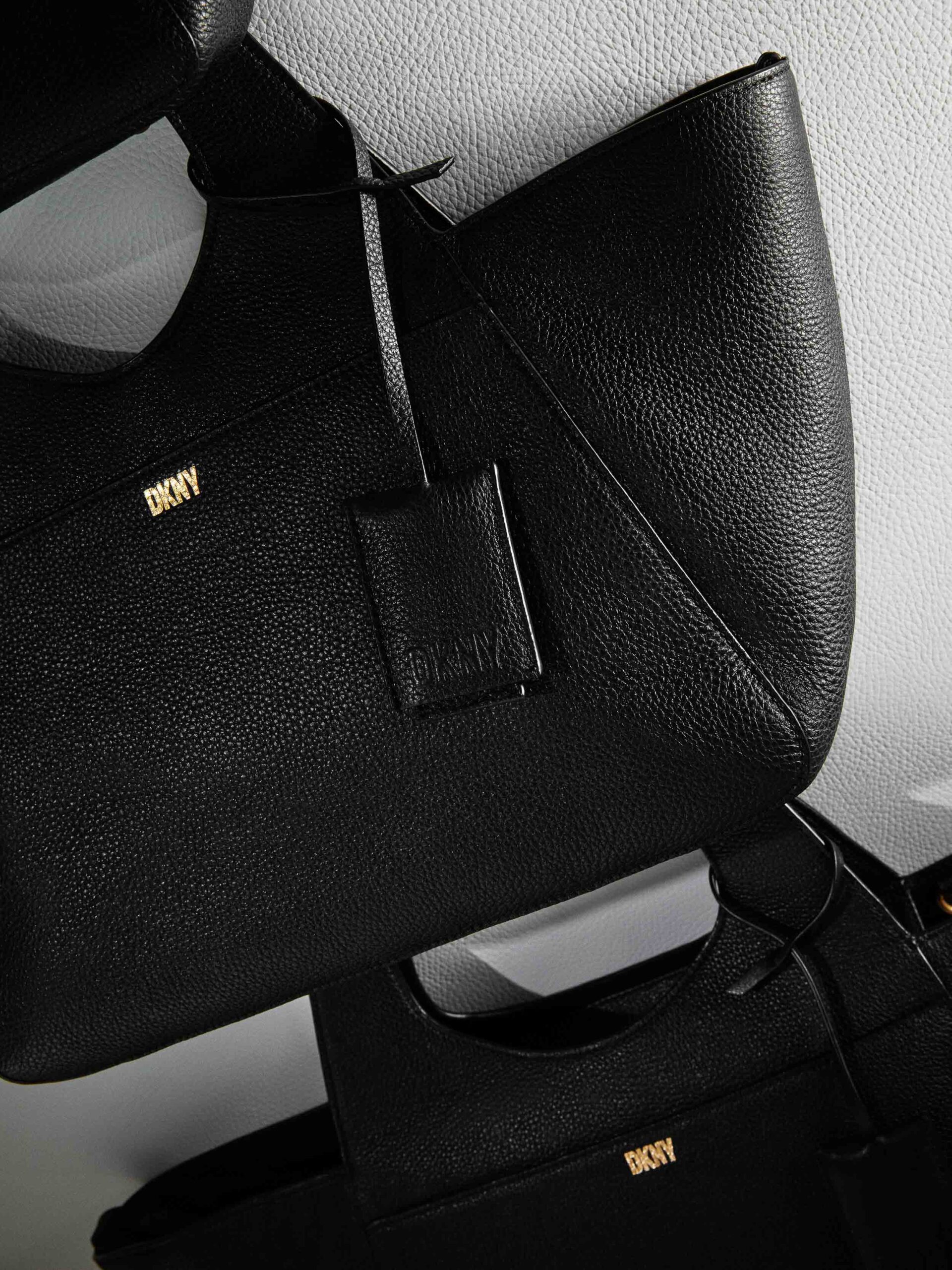Black pebbled leather tote bags with gold logo and leather hang tag, stacked against a white wall, for DKNY.