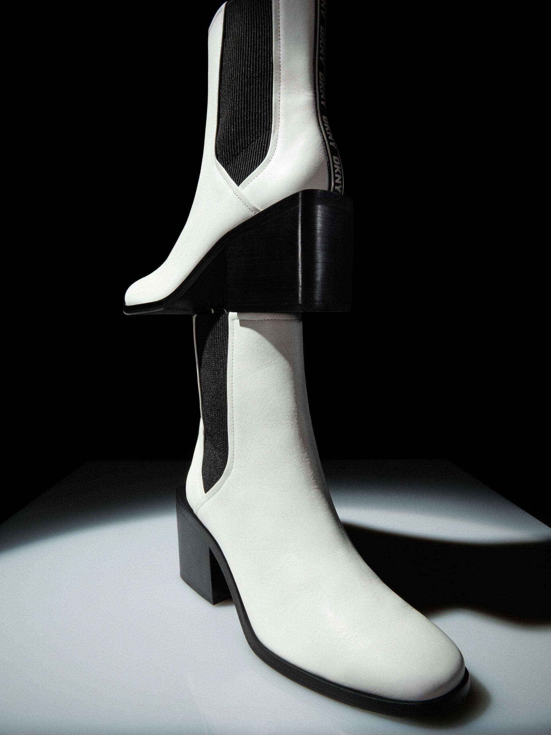Two white leather Chelsea boots with black elastic and block heels stacked on a lit plinth against black, for DKNY.