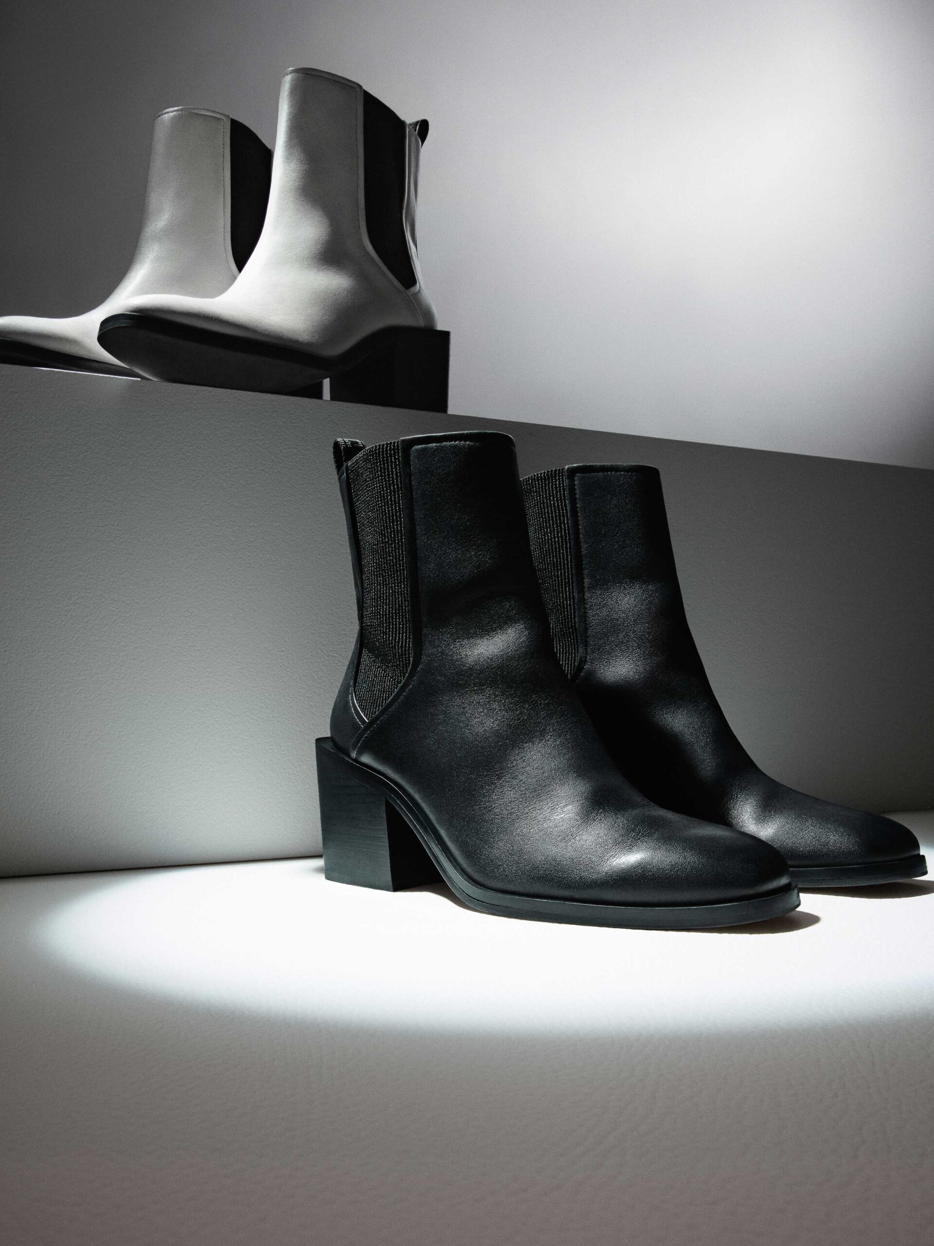 Black leather Chelsea boots in a spotlight below a white pair on a grey step, still life, for DKNY.