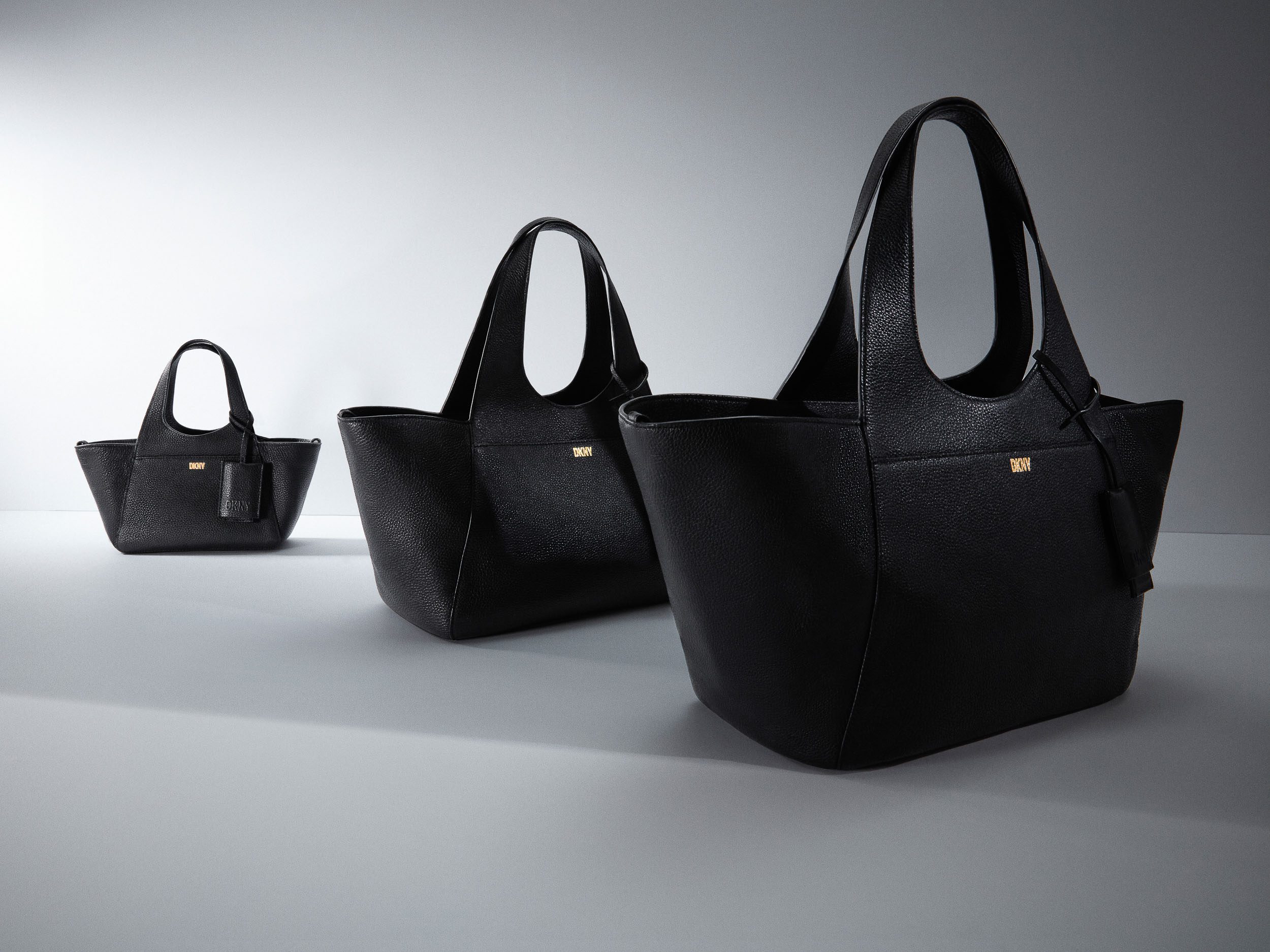 Three black pebbled leather tote bags in graduated sizes with gold logo plates on a grey studio floor, for DKNY.