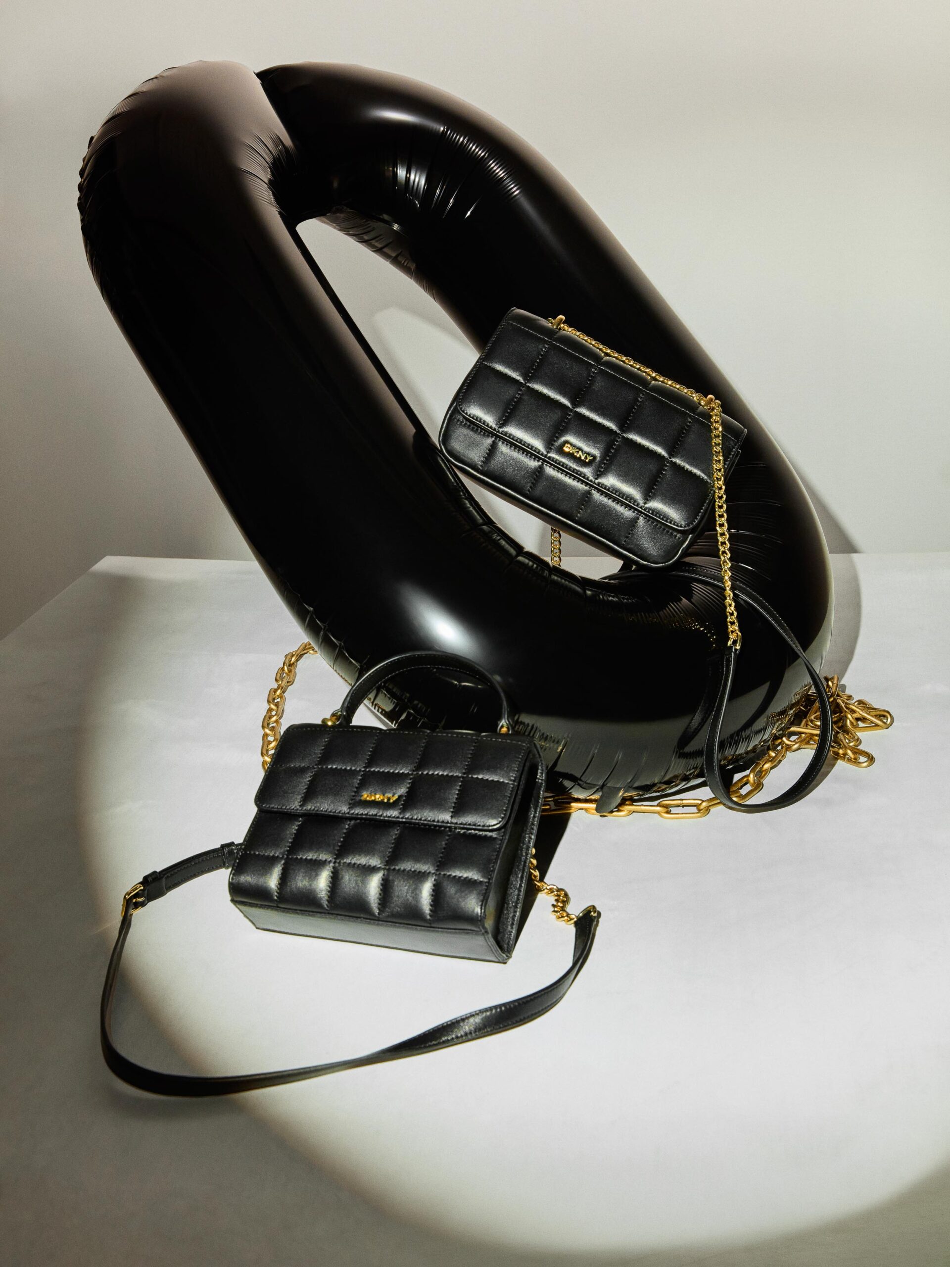 Two quilted black leather bags with gold chain straps on a glossy black inflatable loop in a spotlight, for DKNY.