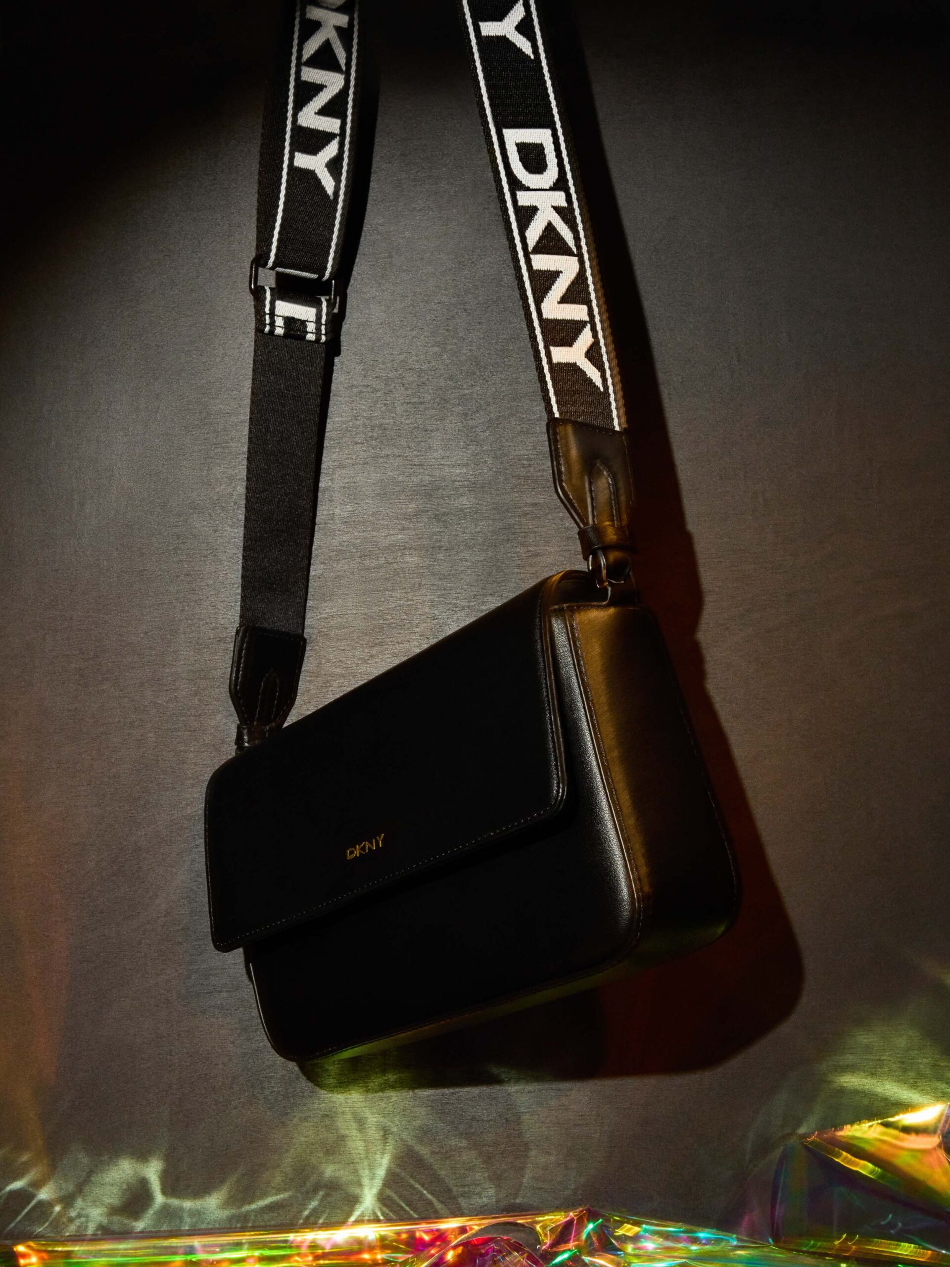 Black leather crossbody bag on a black and white DKNY logo strap, lit over iridescent film on a grey backdrop, for DKNY.